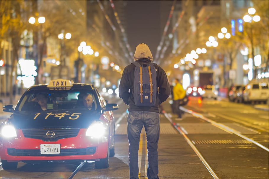 man wearing hoodie and backpack standing on middle of road, taxi, cars, people, backpack, hoodie, jacket, jeans, pants, street, road, city, night, evening, lamp posts, light, urban, driving, HD wallpaper